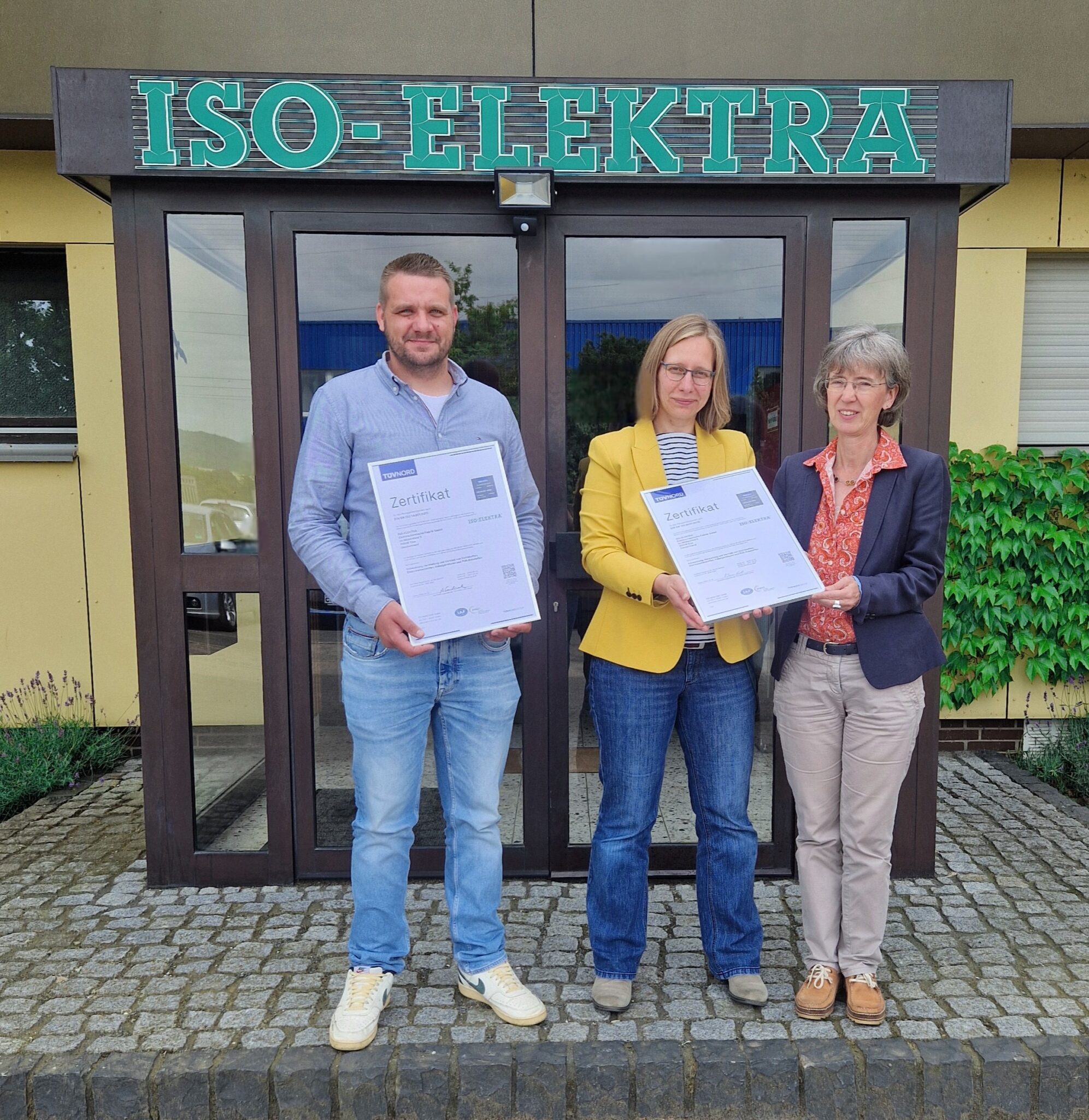 Handover of TÜV certificates to two employees in front of the building entrance