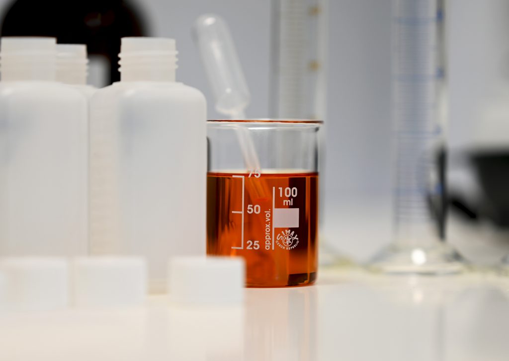 Light brown liquid (additive) in a beaker with pipette. Also measuring cylinder and other bottles.