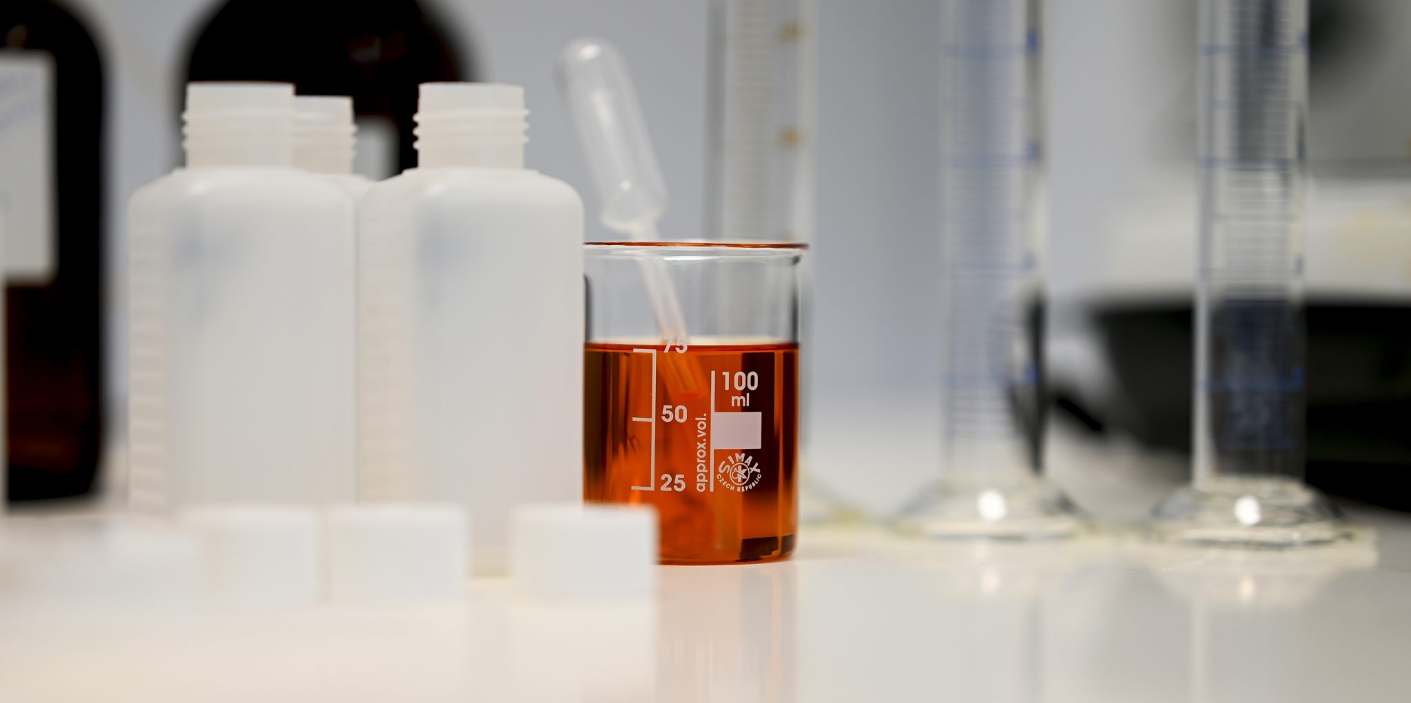 Light brown liquid (additive) in a beaker with pipette. Also measuring cylinder and other bottles.